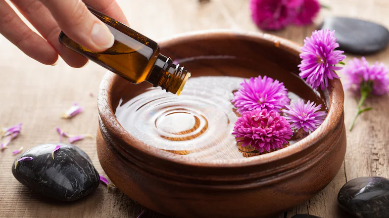 Aromatherapy for stress relief with essential oil being poured into a wooden bowl of water, surrounded by purple flowers and black massage stones for a calming and relaxing experience.